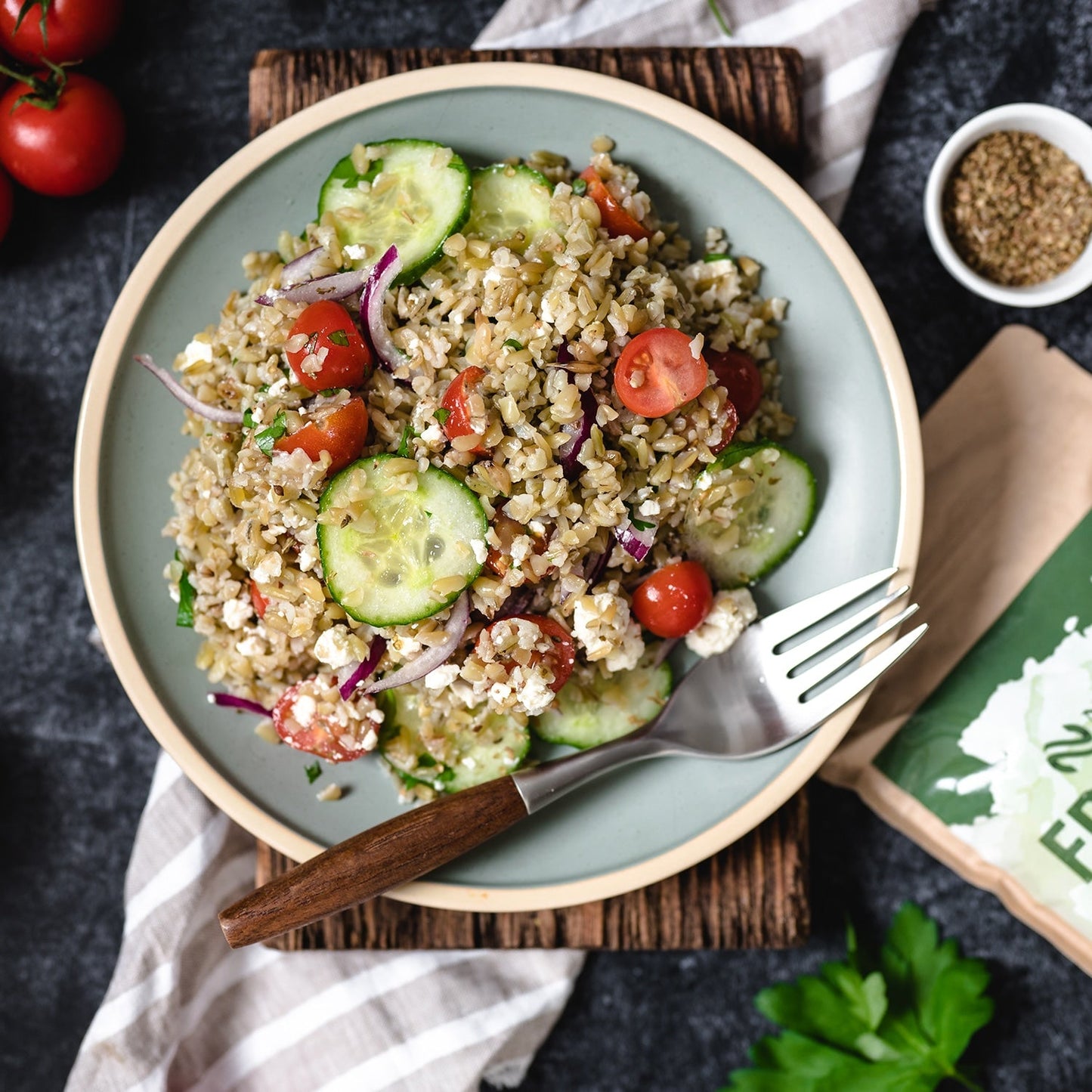 "Ein frischer Freekeh-Salat mit Gurkenscheiben, Kirschtomaten, roten Zwiebeln und Feta-Käse, auf einem rustikalen Holzbrett serviert."