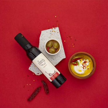 "Eine Flasche Green Gold Olivenöl aus Palästina mit Chili, arrangiert mit einer Schale Oliven, einer Schale mit Dip und Chilischoten auf einem roten Hintergrund."