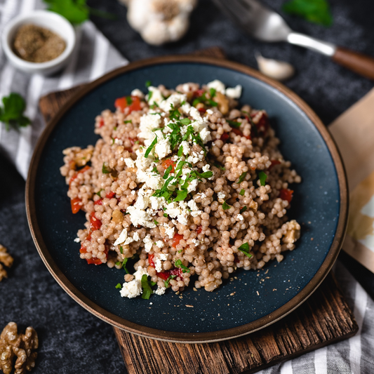Ein Teller mit mediterranem Couscous-Salat, garniert mit zerbröseltem Feta und frischen Kräutern. Im Hintergrund sind Walnüsse und eine Gabel zu sehen.