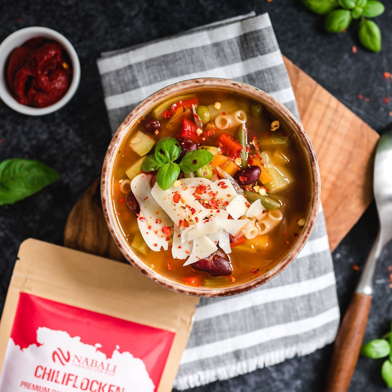 "Eine schmackhafte Gemüsesuppe, garniert mit Chiliflocken und frischen Kräutern, serviert in einer rustikalen Schüssel auf einem Holzbrett."